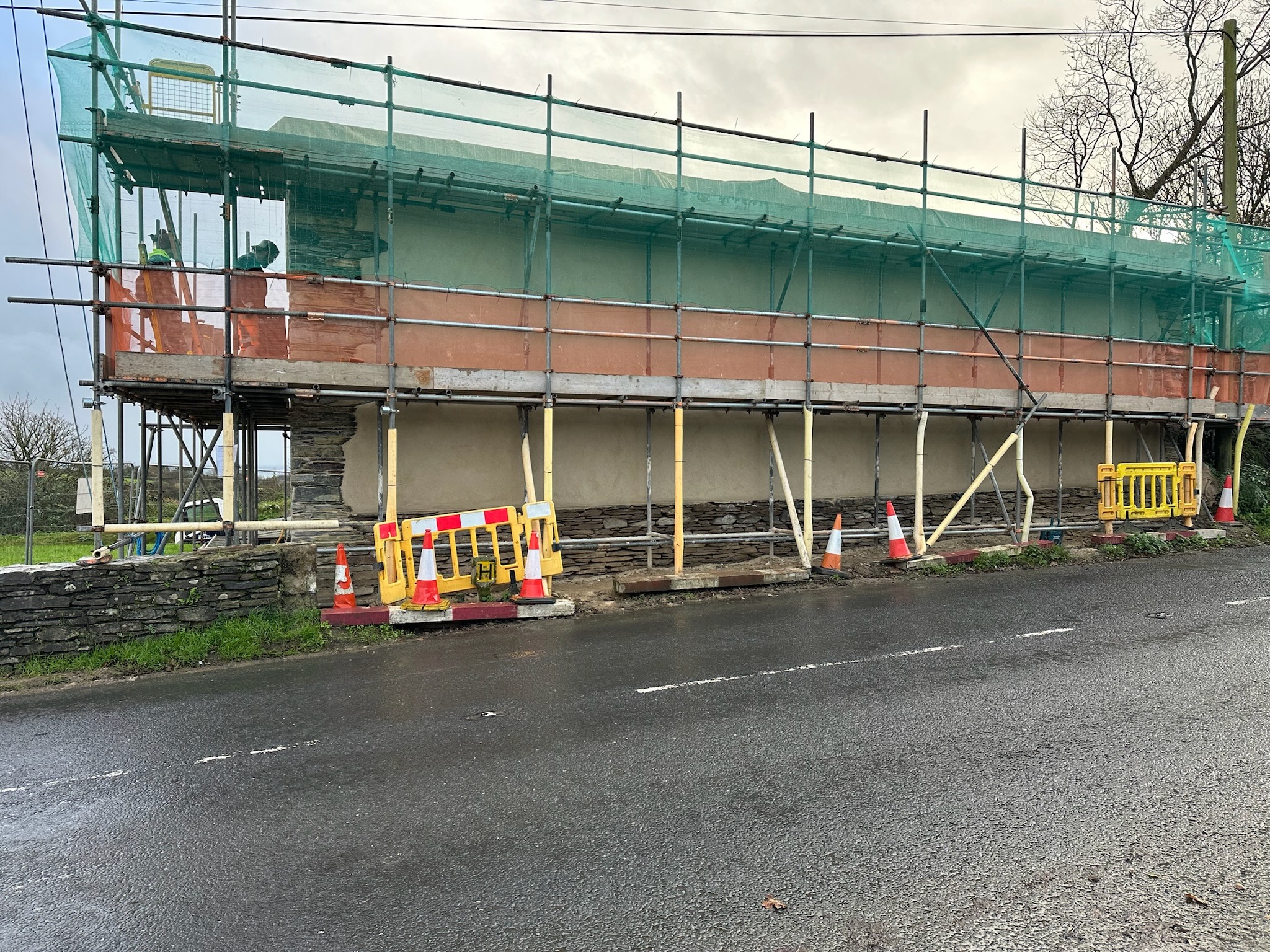 The rebuilt cob wall standing under scaffold during the works, seen from the road